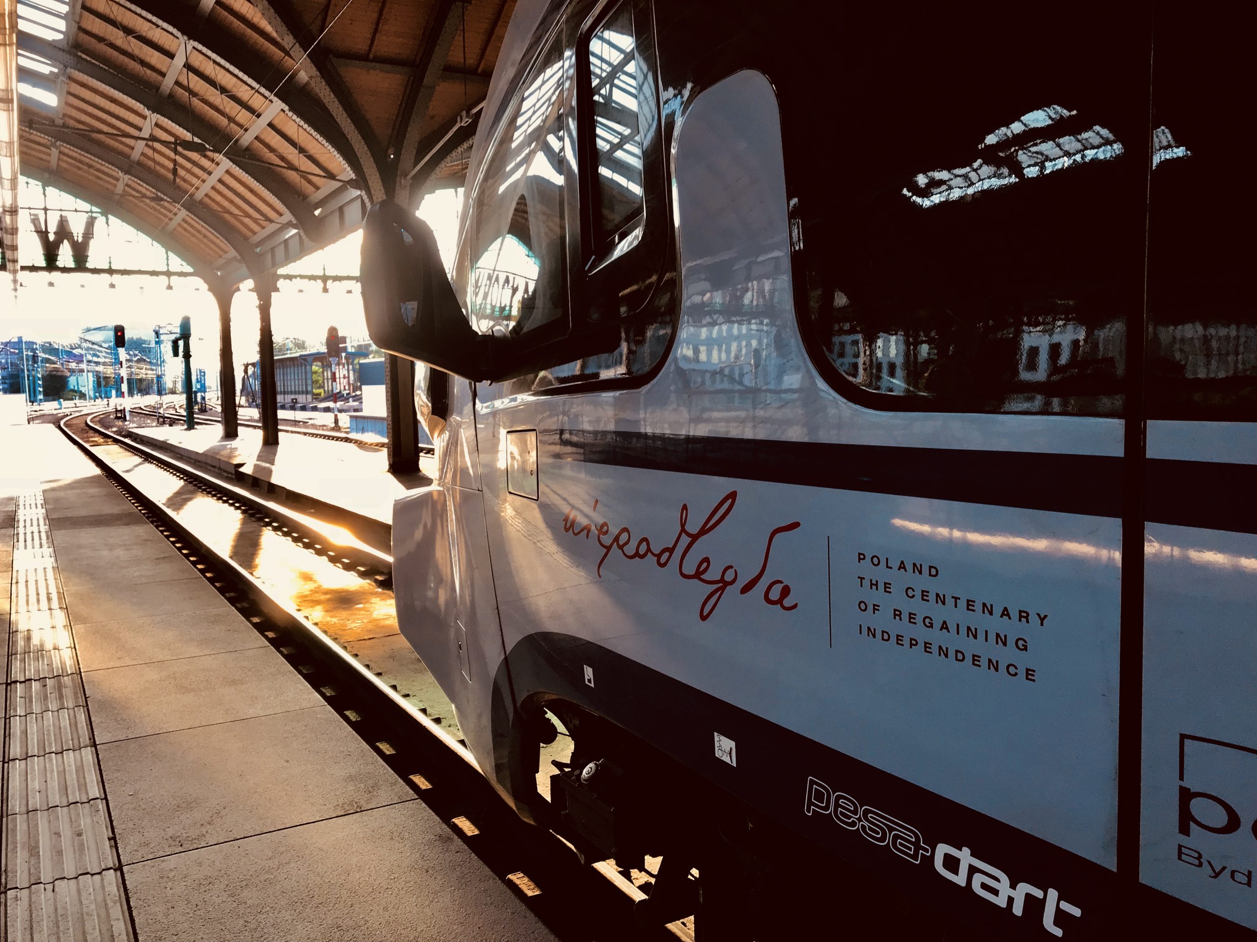 The front of a train engine showing the “Niepodległa” (“Independent”) logo. The train stands in the station against the background of the setting sun