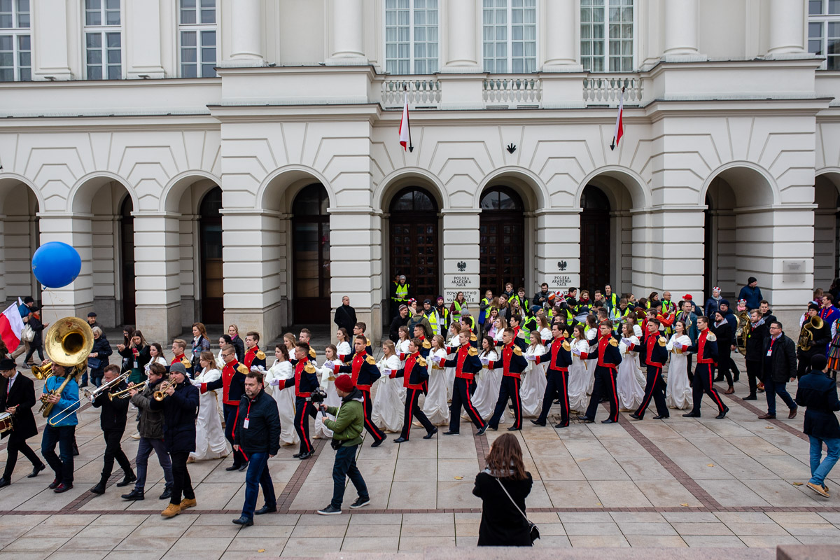 A group of people in festive costumes dancing the polonaise in pairs in the street, accompanied by a band and watched by passers-by