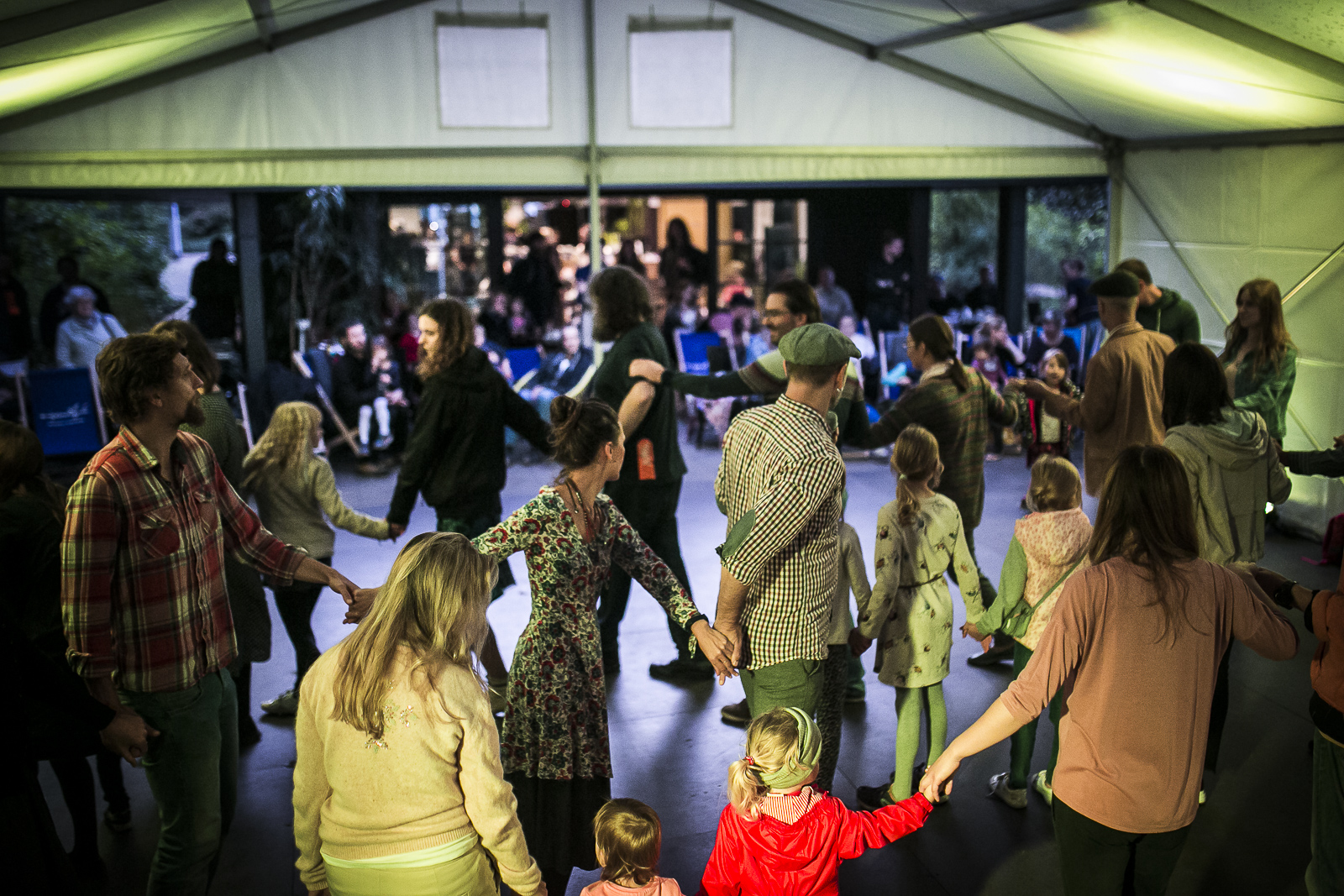 A photograph showing people dancing under a tent. Participants, including children, dance in a circle holding hands; in the background, there is a pavilion and spectators sitting on deck-chairs