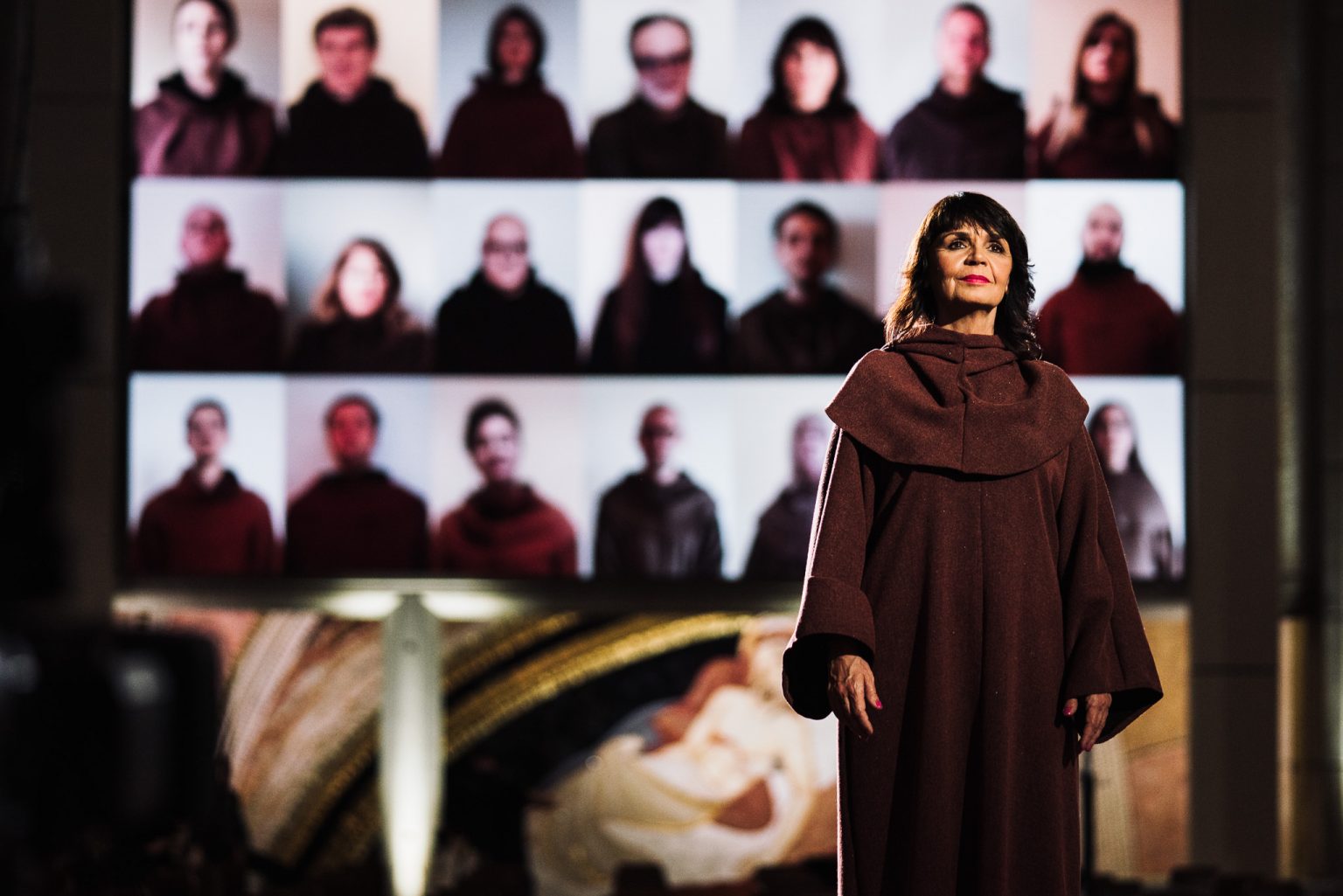 In the foreground, a female performer in a modest dress reminiscent of a nun’s frock; in the background, a screen showing members of the choir 