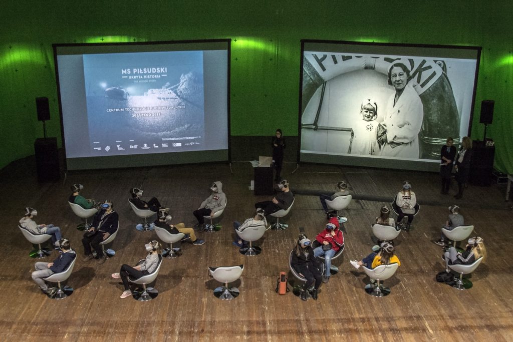 A colour photograph showing people in virtual goggles watching a VR movie. In the background, a still from the film and archival photos from the deck of the MS Piłsudski are displayed on screens.