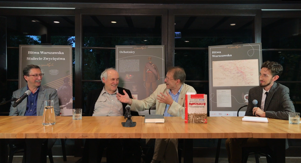 Three older men talking at a table, smiling. On the right, a younger man – the facilitator of the debate. In the background, boards from an exhibition dedicated to the 1920 Battle of Warsaw