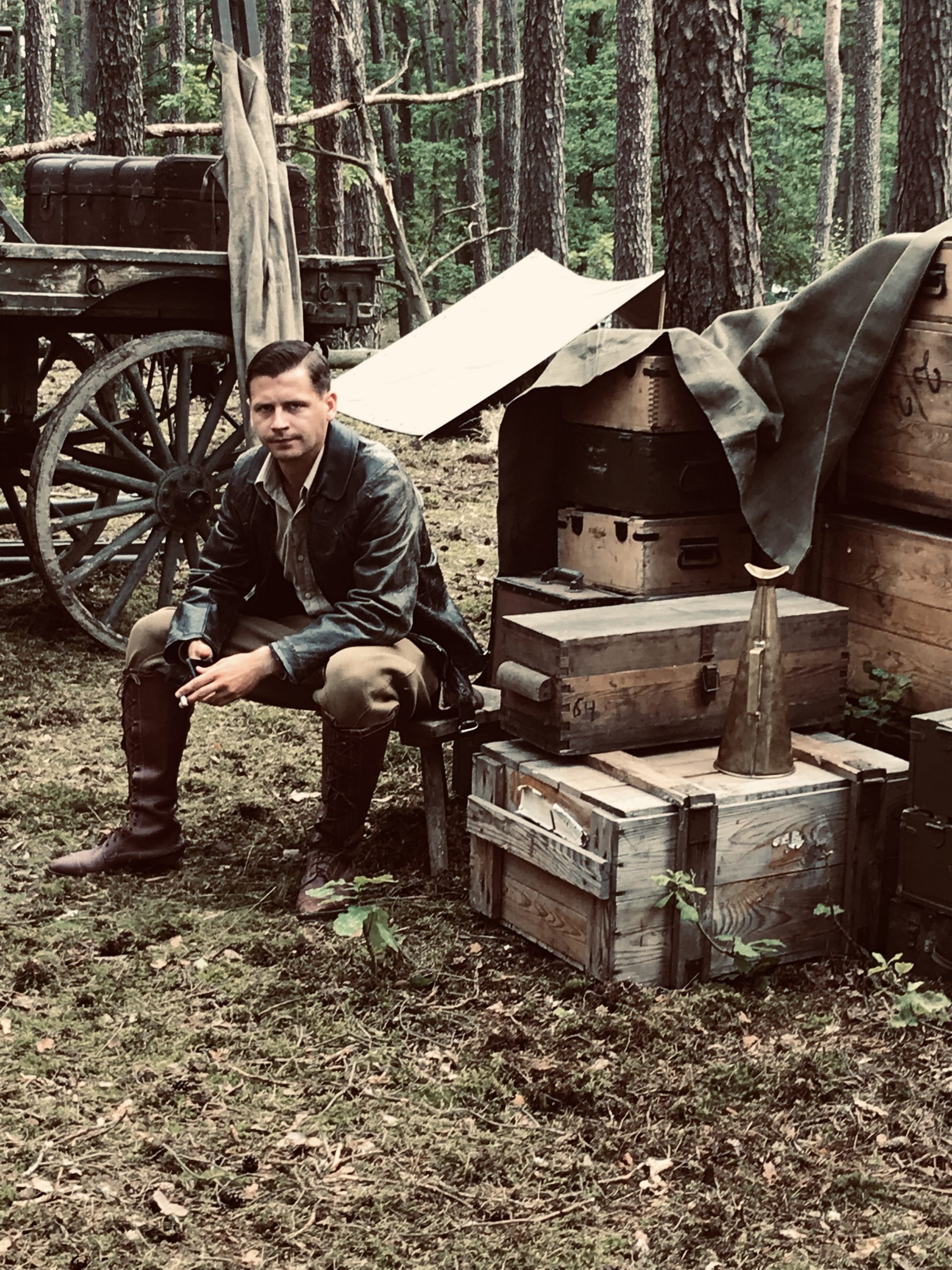 Description: A man in an early 20th century costume sitting in a forest on a stool next to stacked chests; in the background, part of a wagon is visible
