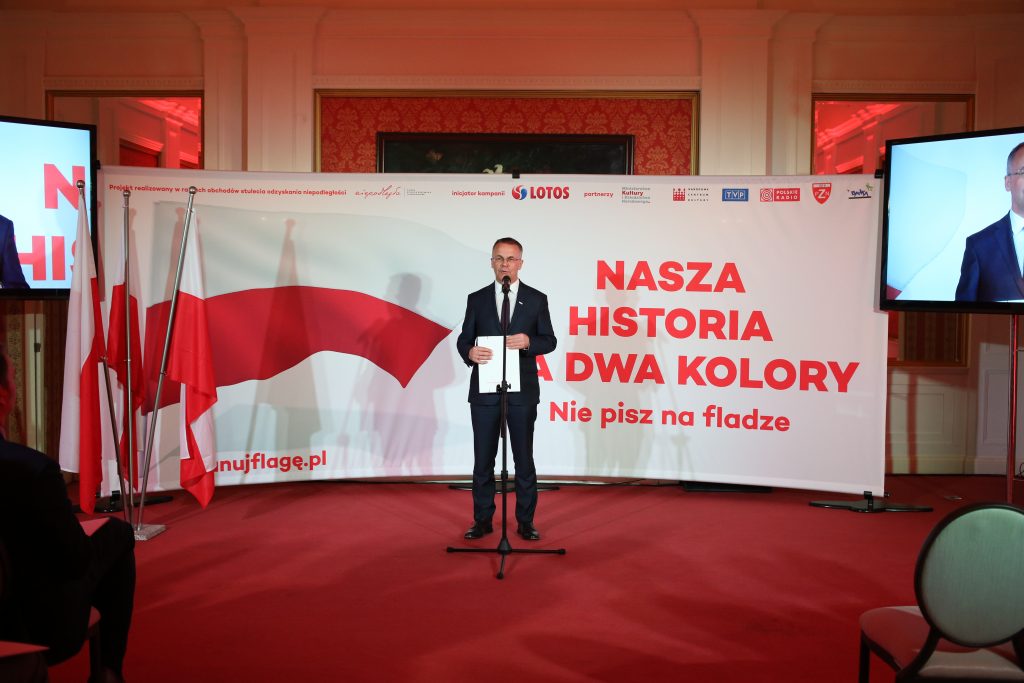 mężczyzna elegancko ubrany stoi na środku przed mikrofonem, w tle baner z flagą Polski i hasłem kampanii