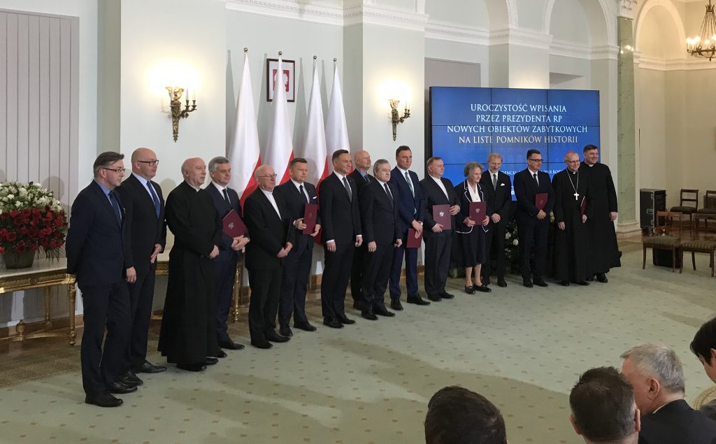 w rzędzie stoją elegancko ubrani opiekunowie zabytków, trzymają teczki z rozporządzeniami, w środku prezydent, w tle flagi Polski i ekran