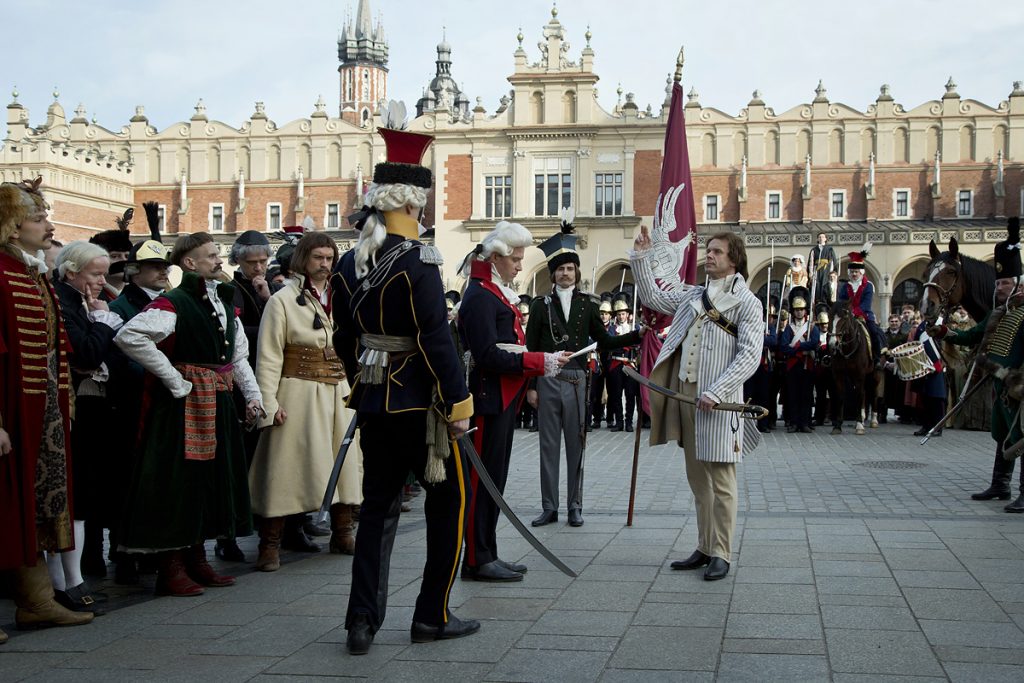 Kościuszko stoi na środku w postawie uroczystej z uniesioną do ślubowania ręką, wokół zgromadzeni gapie, w tle sukiennice krakowskie