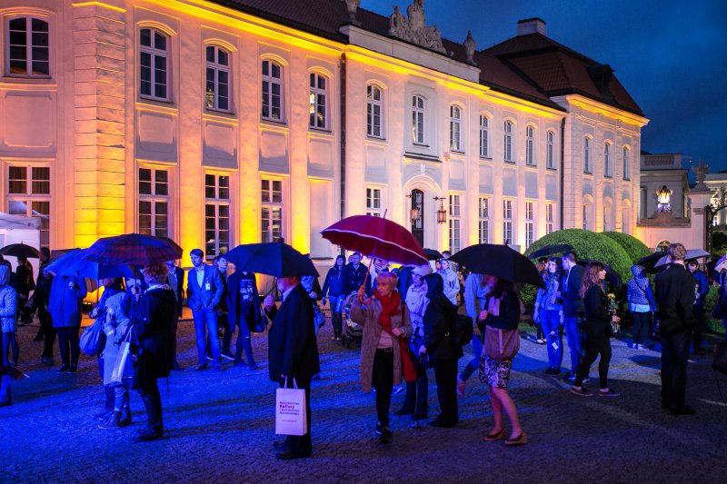 grupa ludzi stoi przed kolorowo oświetlonym pałacem, w rękach mają parasolki i torby, kręcą się i oglądają