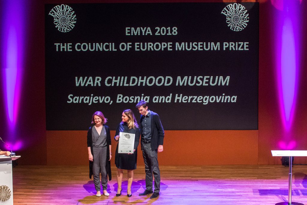 troje ludzi na podium, prezentują odbieraną nagrodę, w tle napis opisujący muzeum