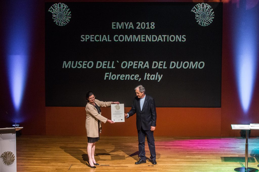 dwoje ludzi na podium, prezentują odbieraną nagrodę, w tle napis opisujący muzeum