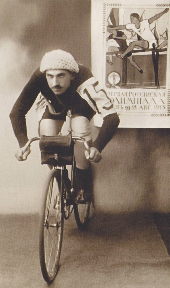 kolarz z wąsem i w kasku na rowerze w studiu fotograficznym, z plakatem olimpiady na ścianie