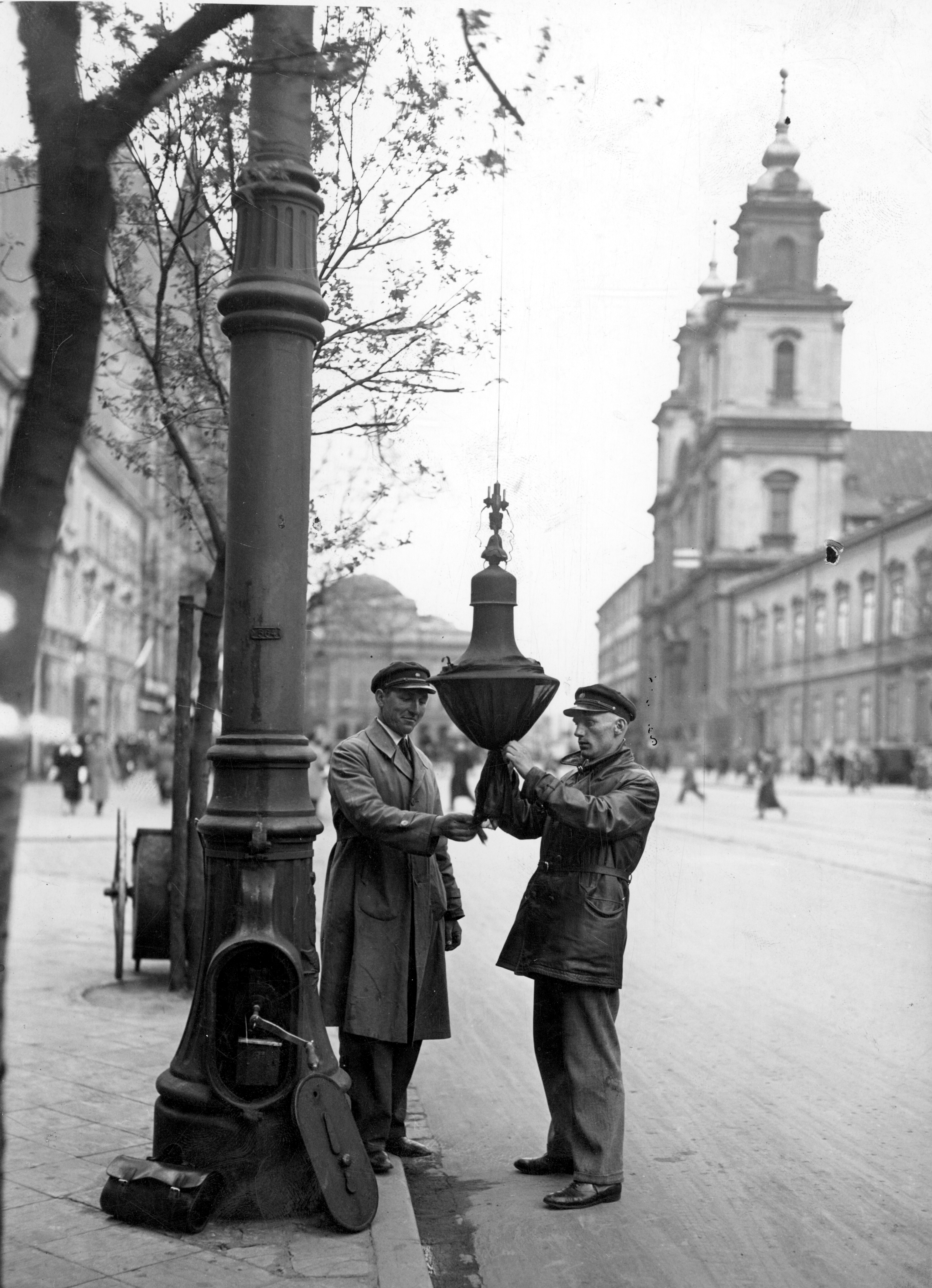 Dwóch mężczyzn zawiesza dekoracje żałobne na latarniach przy Krakowskim Przedmieściu.
