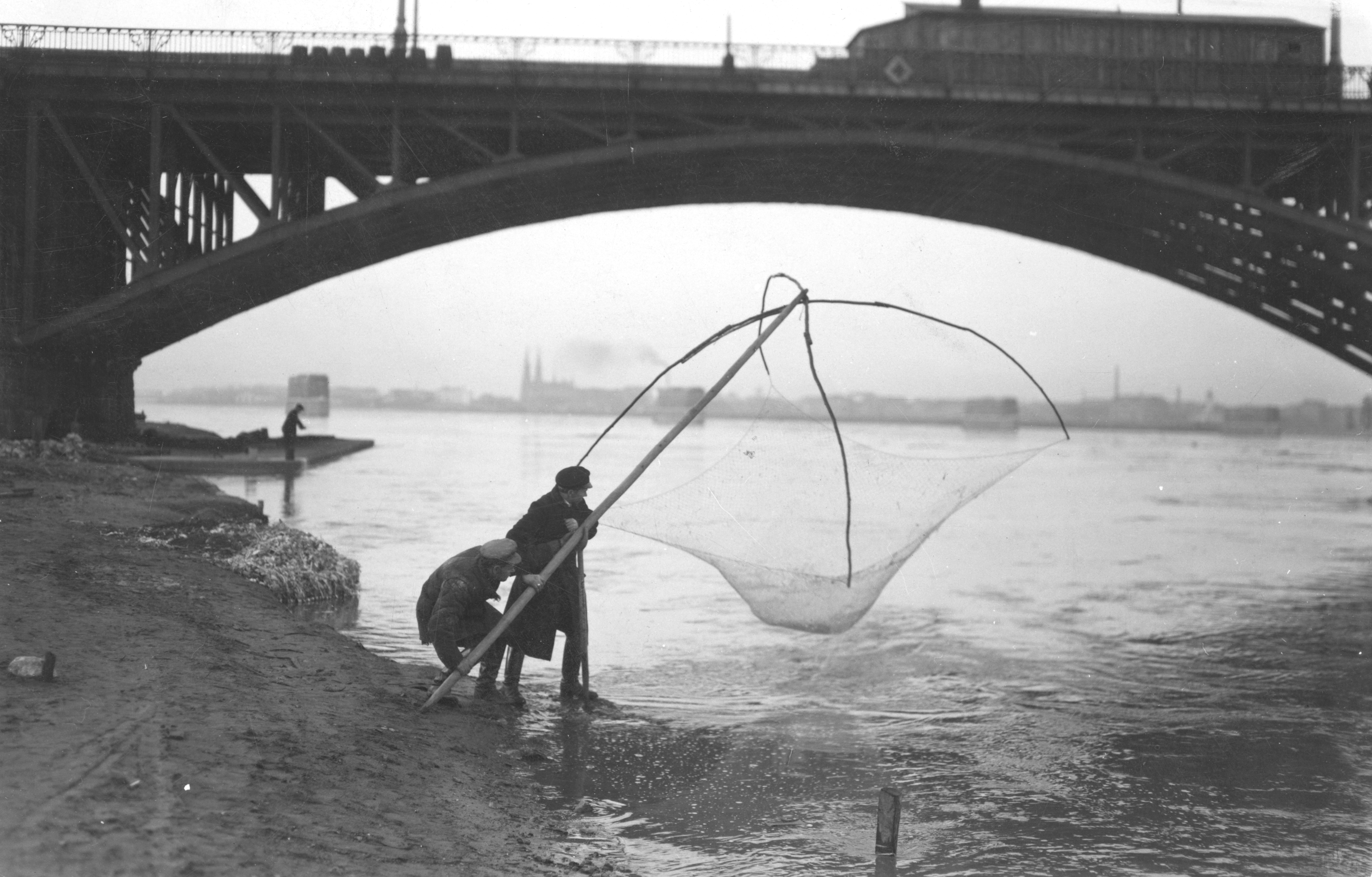 Dwóch mężczyzn stoi z podrywką na brzegu Wisły w Warszawie. Widoczny fragment przęsła mostu Poniatowskiego oraz fragment praskiego brzegu Wisły. 
