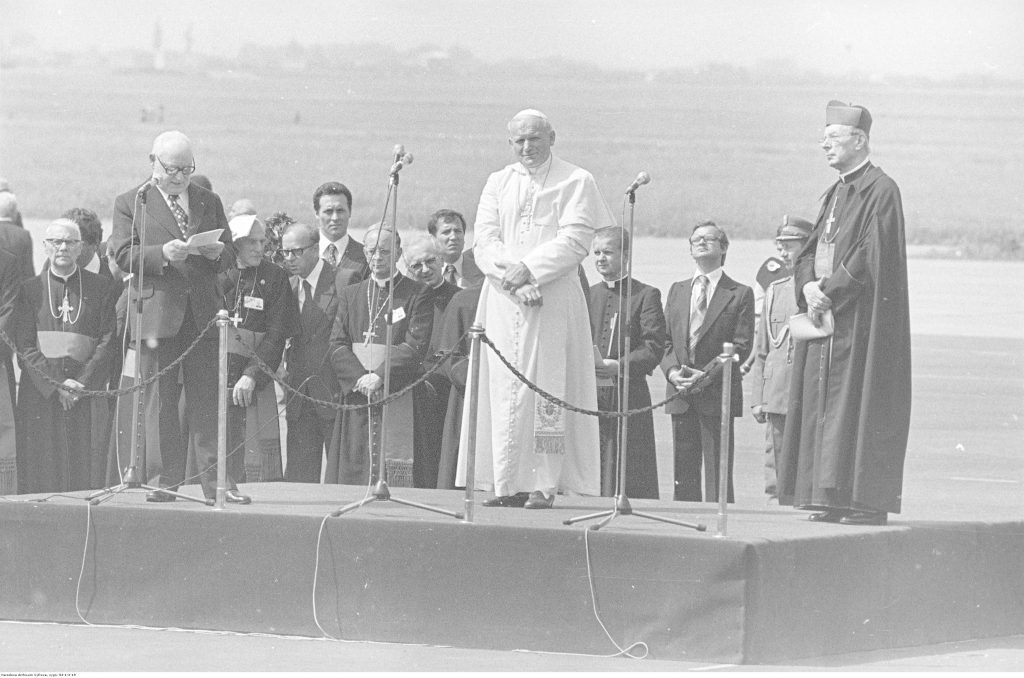 na lotnisku ustwione podium, na nim stoją papież i inny przemawiający mężczyzna, w tle rząd mężczyzn w odświętnych strojach