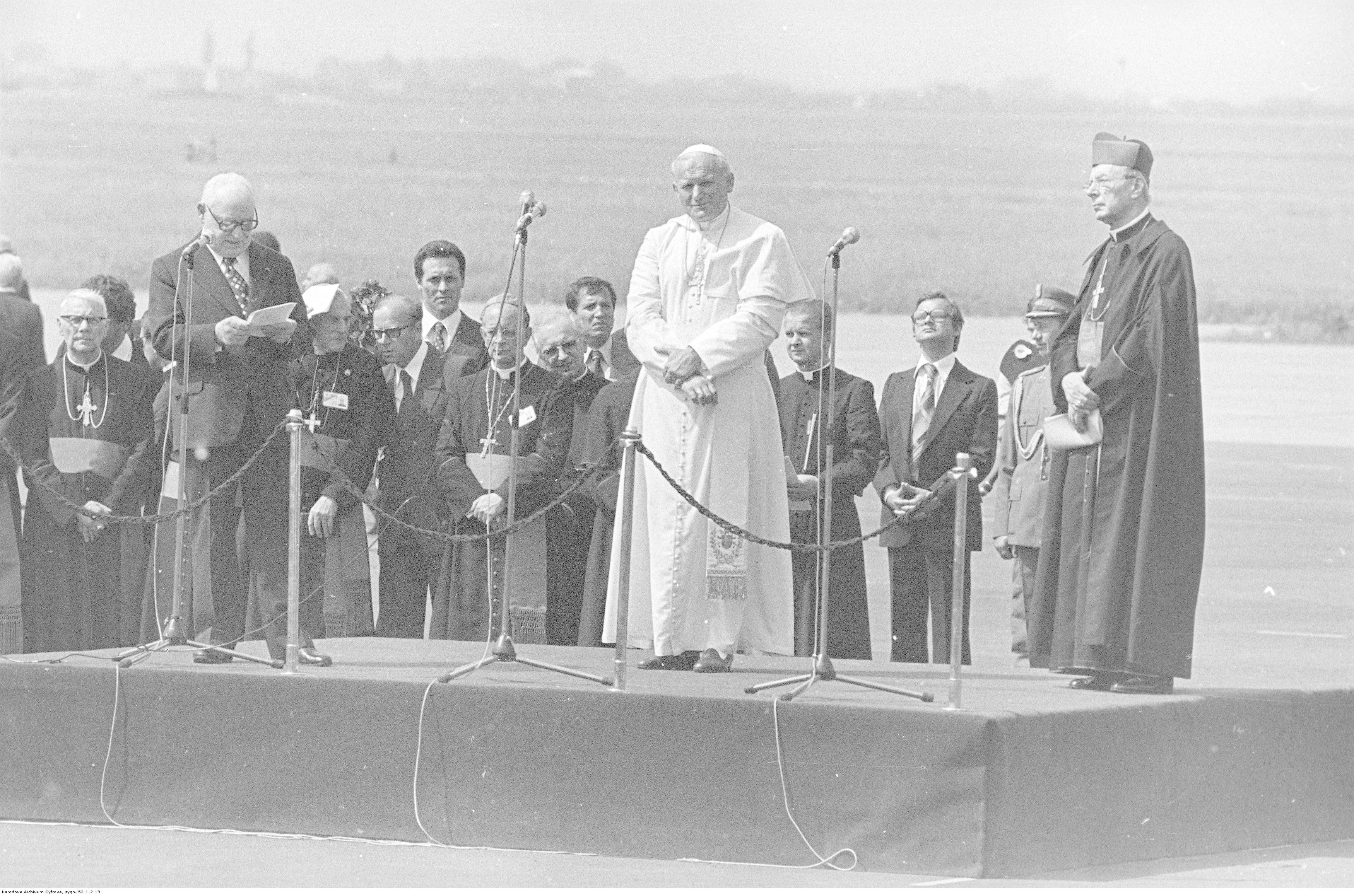 na lotnisku ustwione podium, na nim stoją papież i inny przemawiający mężczyzna, w tle rząd mężczyzn w odświętnych strojach