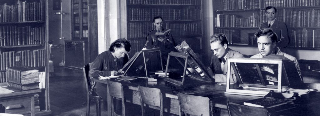 pracownicy w bibliotece przy biurku czytają stare, wielkie księgi