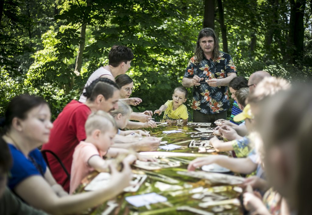 dzieci z rodzicami przy stole podczas warsztatów manualnych, w tle prowadzący