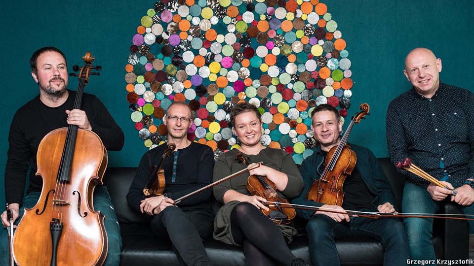 5 muzyków z instrumentami siedzi na tle ściany z okrągłą dekoracją