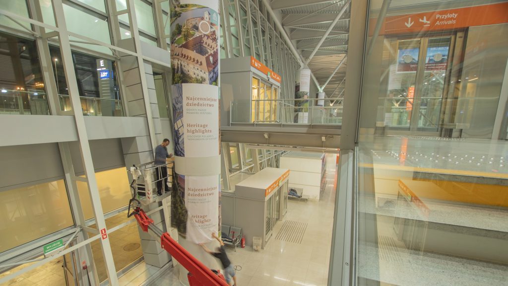 zdjęcie przedstawia mężczyznę montującego wystawę na filarach w hali portu lotniczego na Okęciu