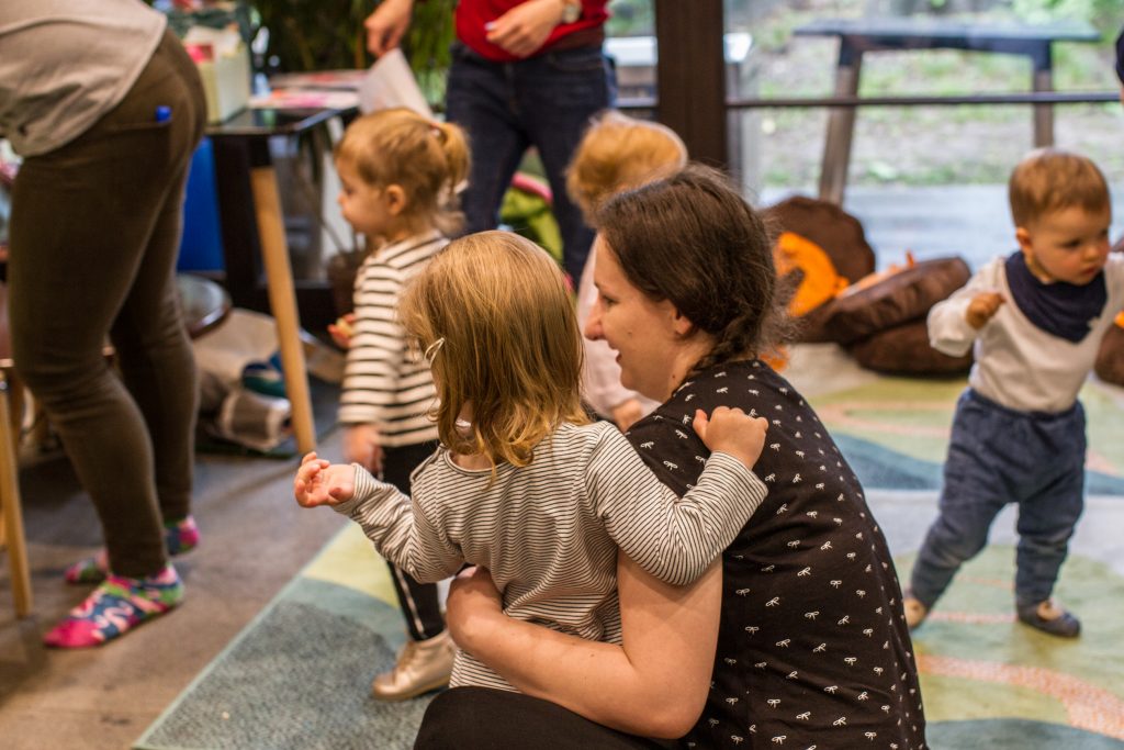 zdjęcie przedstawia kobietę przytulającą dziecko i dzieci biegające wokoło - ilustracja obrazująca warsztaty sensoryczne dla dzieci