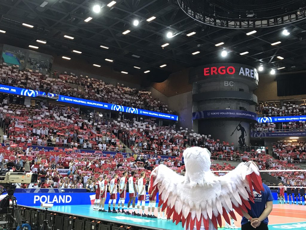 Kibice na trybunach podczas śwpiewania hymnu z uniesionymi kartkami kartoniady, na pierwszym planie odwrócona tyłem maskotka reprezentacji - orzeł biały z uniesionymi skrzydłami