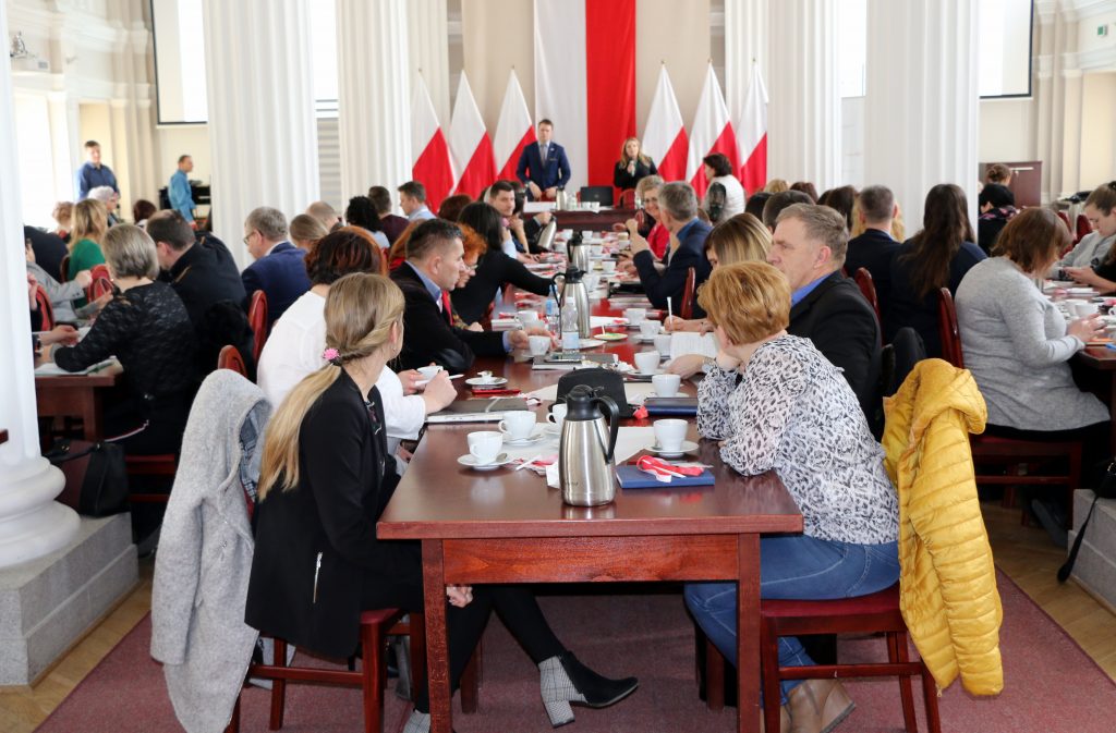 grupa ludzi siedzących przy trzech równoległych stołach, w tle stojące postaci w garniturach, które prowadzą spotkanie