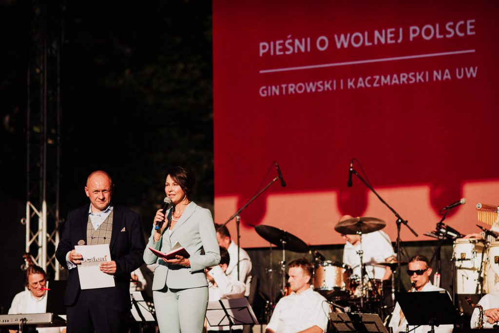 dwoje prowadzących na scenie, za nimi muzycjy, m.in. perkusja, w tle czerwona płachta z nazwą koncertu