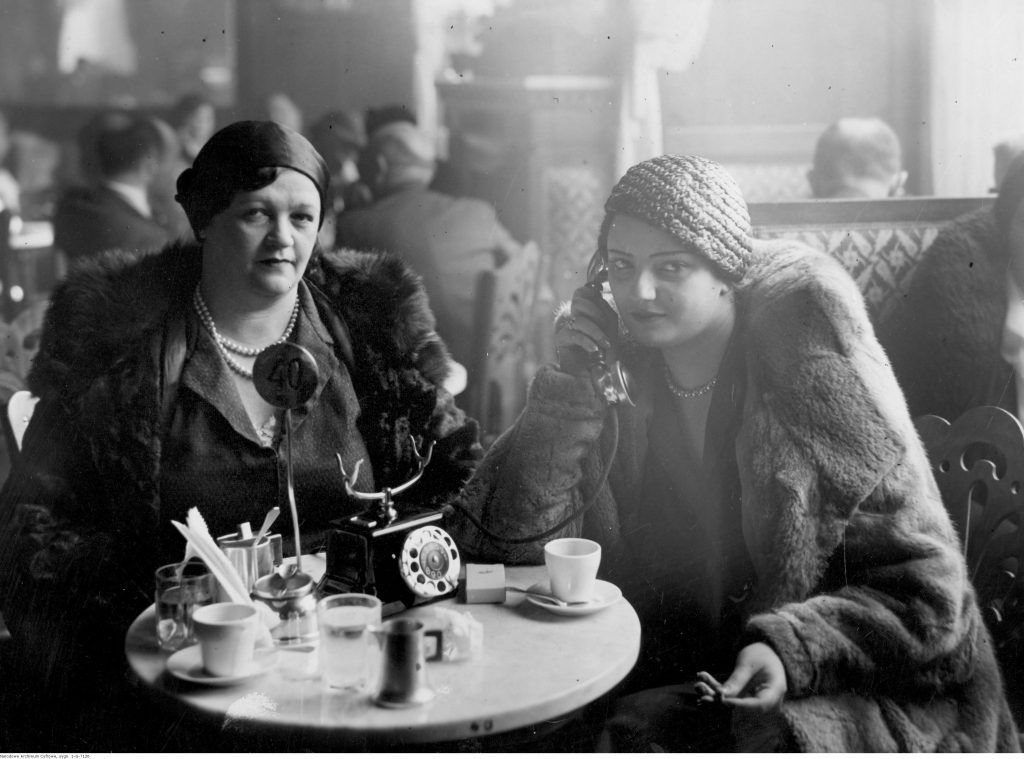 Goście restauracji siedzący przy stolikach. Na pierwszym planie widoczne dwie kobiety, jedna z nich rozmawia przez telefon stojący na stoliku.