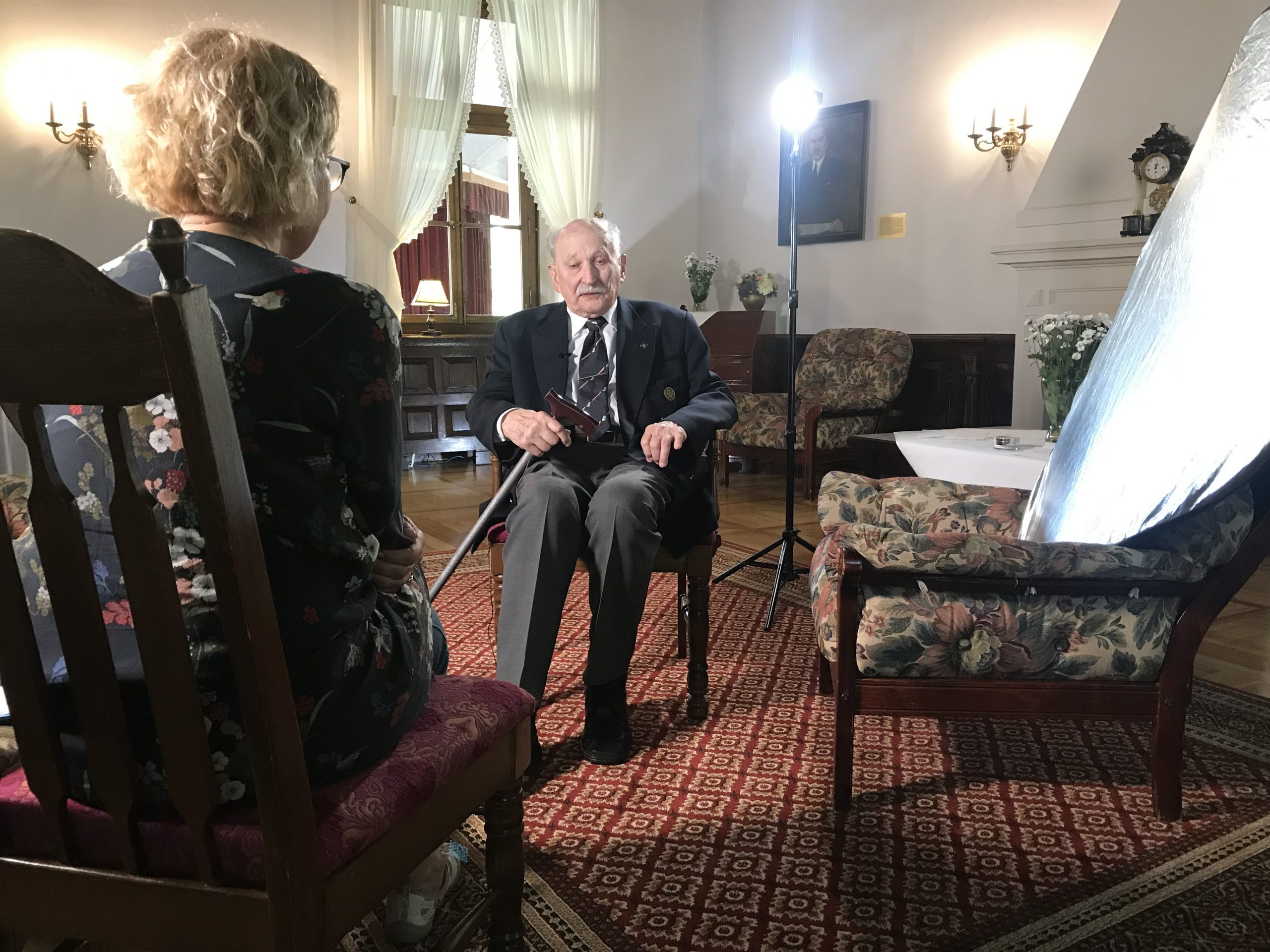 zdjęcie przedstawia plan nagraniowy - na pierwszym planie kobieta siedząca tyłem, na środku zdjęcia starszy mężczyzna siedzący na krześle