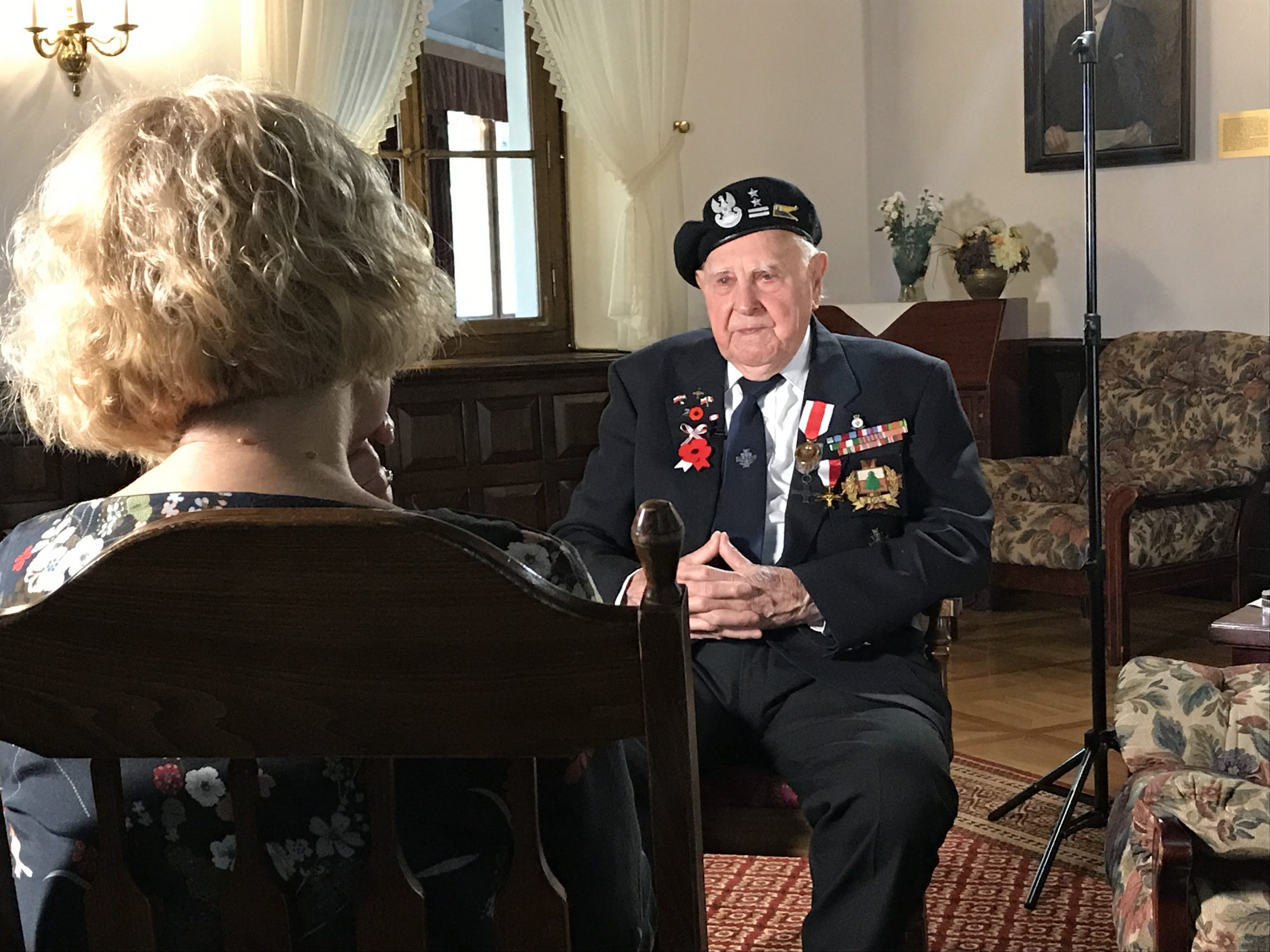 zdjęcie przedstawia plan nagraniowy - na pierwszym planie kobieta siedząca tyłem, na środku zdjęcia starszy mężczyzna siedzący na krześle