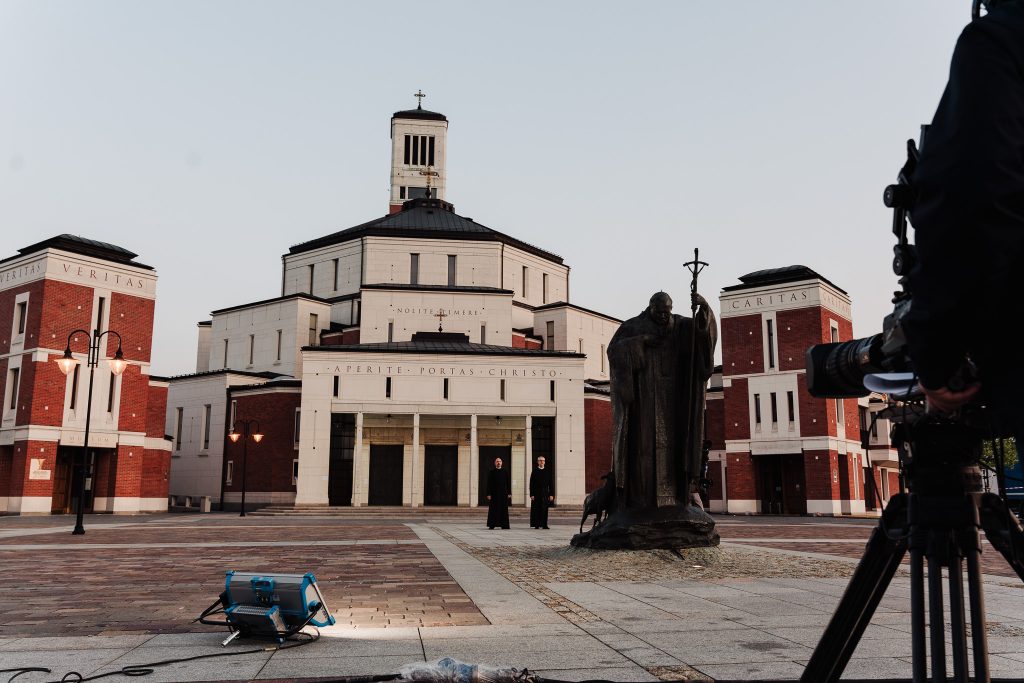 budynek sanktuarium JP II, na pierwszym planie kamera, dalej w centrum pomnik JPII, a dalej II mnisi przed budynkiem