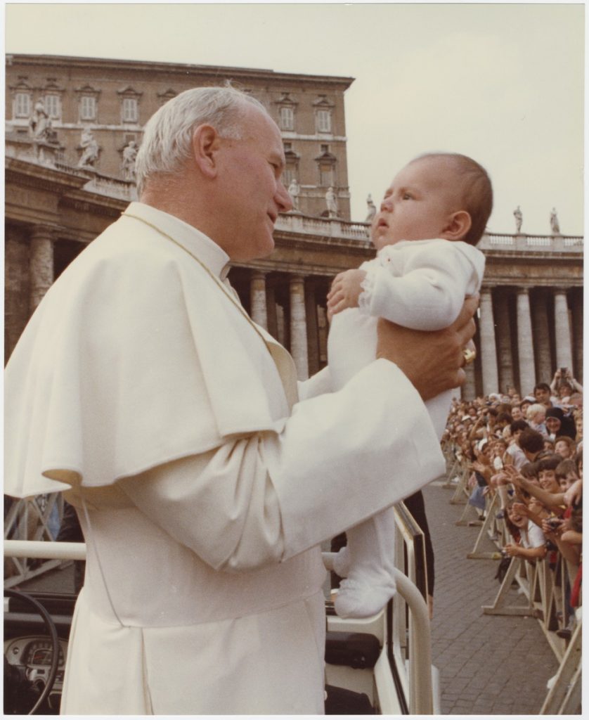 jan paweł II w papamobile trzyma na rękach bobasa, w tle plac św piotra pełen pielgrzymów machających