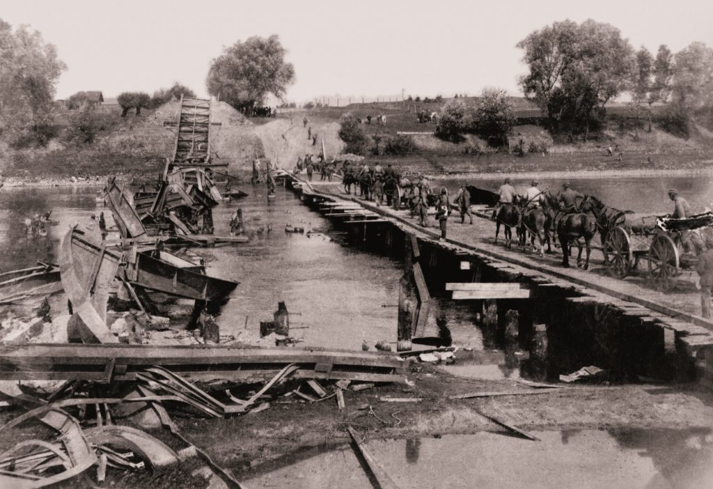 tymczasowy drewniany most, po którym idą konie i wozy, obok zrujnowanego mostu w rzece