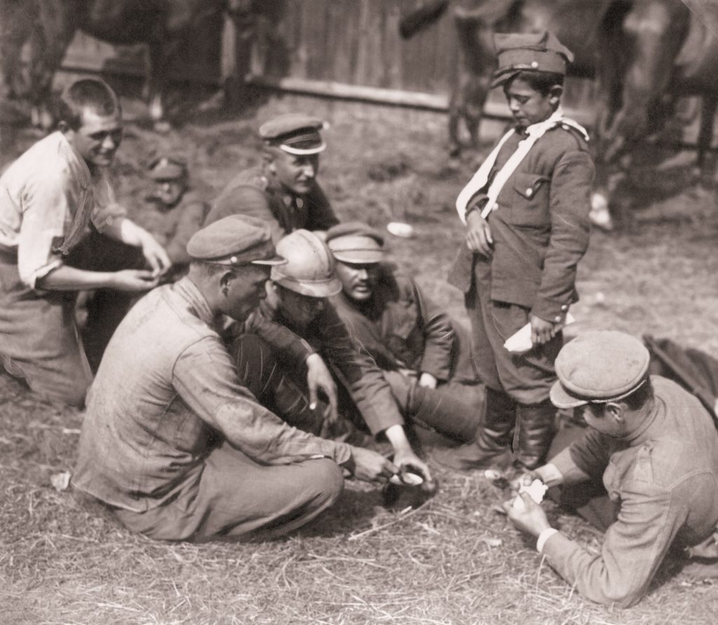 grupa żołnieży jedzących z jednej menażki, przy nich stoi chłopczyk z ręką na temblaku