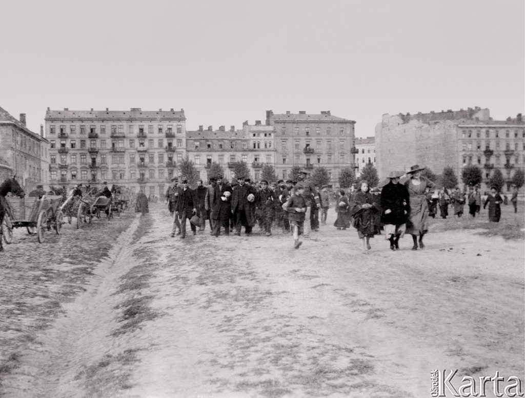 grupa maszerującej ludności, a tle międzywojenne bloki