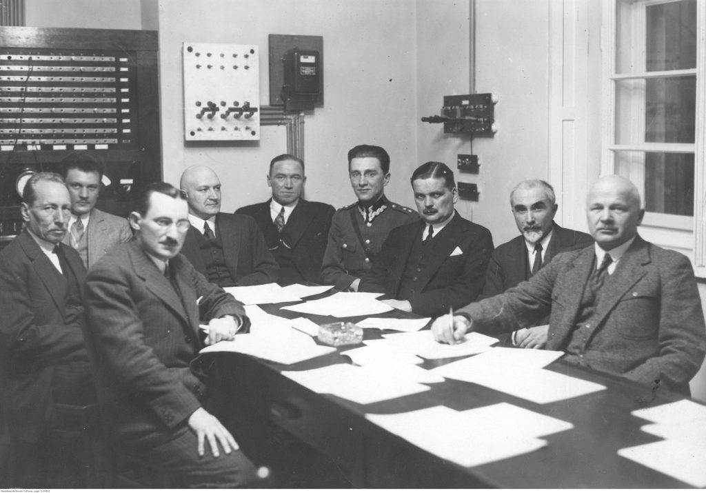 męzcczyźni siedzą przy stole. Widoczni od lewej: prof. Janusz Groszkowski, dyrektor Instytutu prof. Dymitr Sokolcow, wicedyrektor Instytutu W. Cichowicz, sekretarz Roman Rudniewski, płk. Ornbach, inż. S. Jasiński, dyrektor Kazimierz Jackowski, prof. M.Pożaryski, prof. K.Drewnowski.