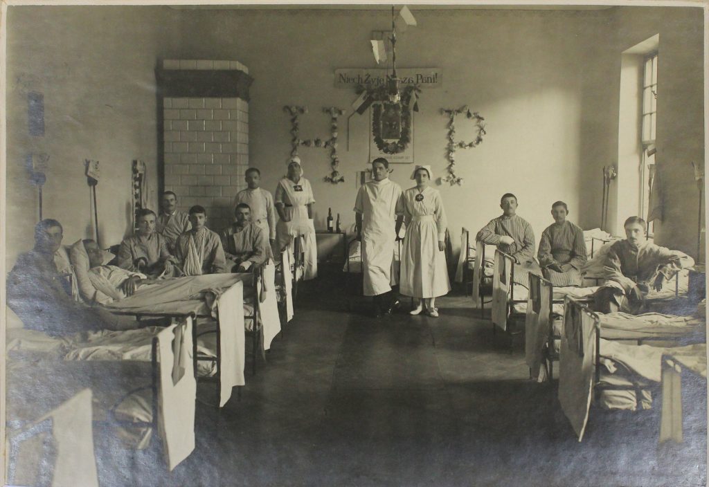 sala szpitalna, na łóżkach pod ścianami siedzą ranni mężczyźni, w centrum stoją 2 sanitariuszki z lekarzem, w tle na ścianie z kwiatów litery H i P