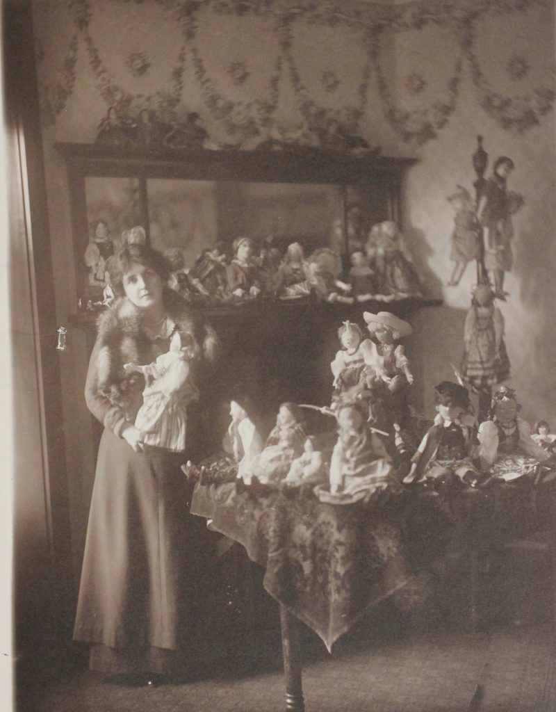 Kobieta w sukni w uksusowym wnętrzu, na stole przed nią i na kredensie za nią poukładane rzesze lalek