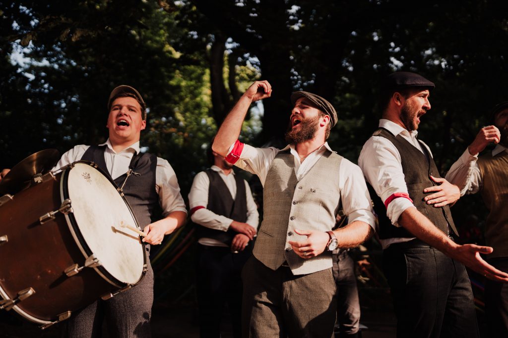 4 mężczyzn w retro ubraniach podczas występu z opaskami AK na rękawach, w tle drzewa