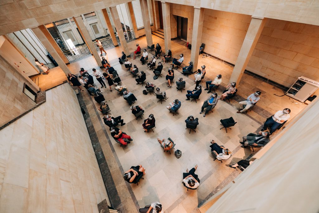 w sali wyłożonej marmurem ludzie sziedzą na brotowych krzesłach w równych odtępach z googlami VR na oczach