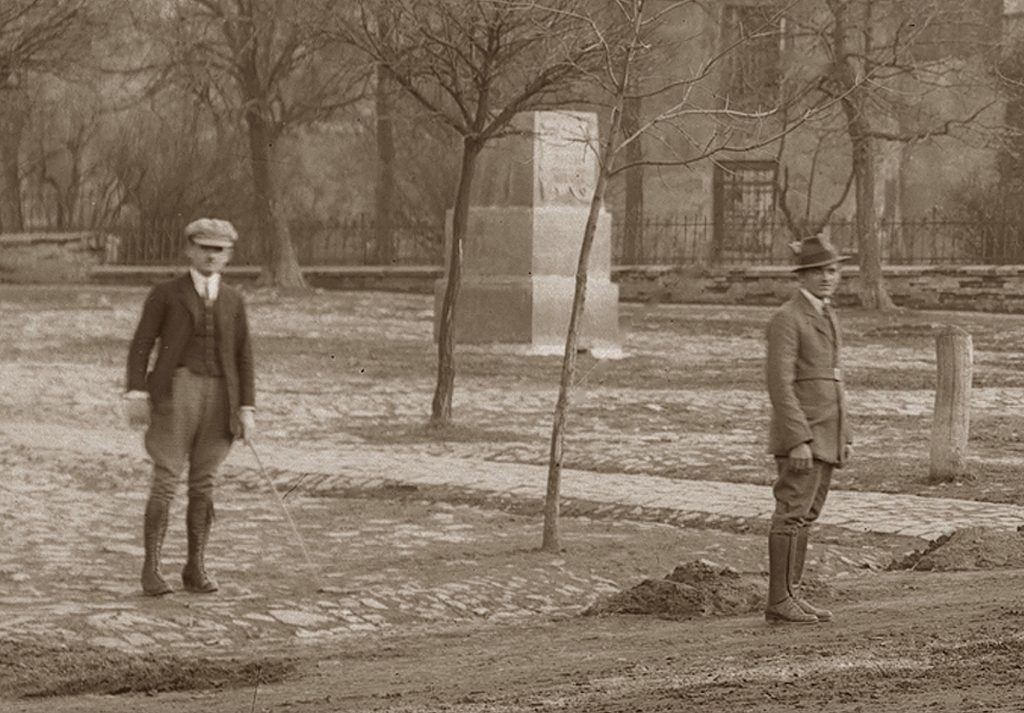 dwaj mężczyźni stojący w parku z młodymi drzewami, za nimi bryła pomnika