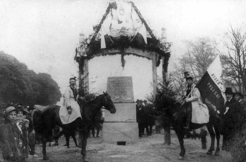 pomnik pod odświętną bramą, obok stoją ułani na koniach