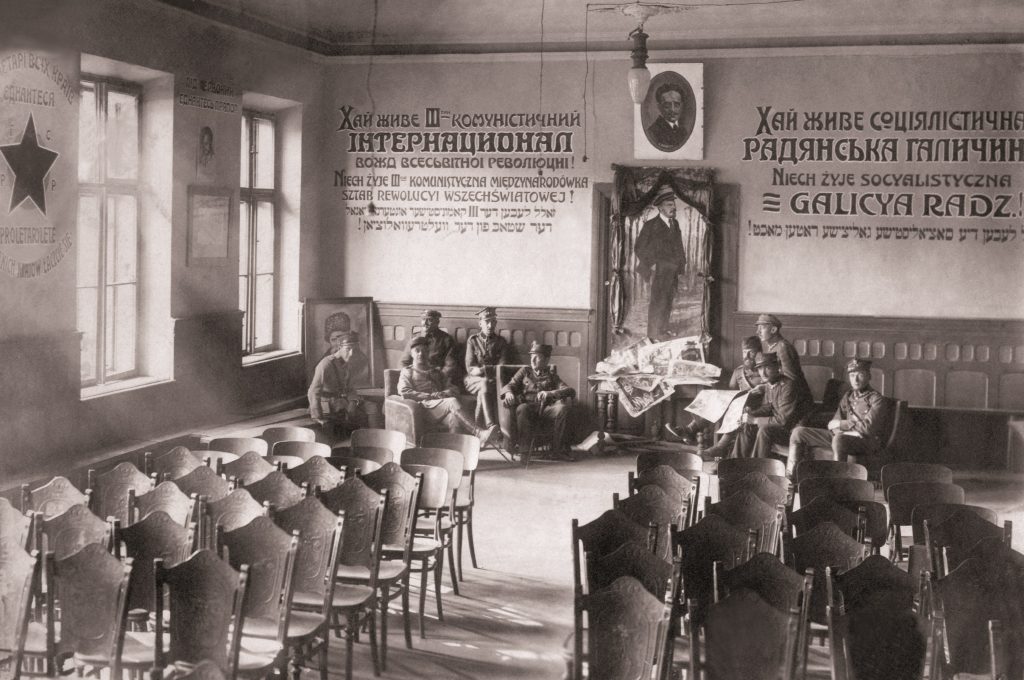 klasa z krzesłami, na ścianach napisy cyrylicą i ozdobione portrety, pod nimi pozuje kilku żołnierzy