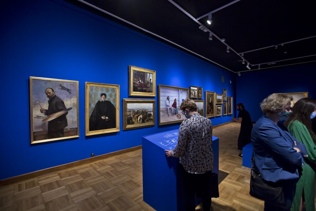 ściana z zawieszonymi obrazami, na pierwszym planie kobieta przy ekspozytorze