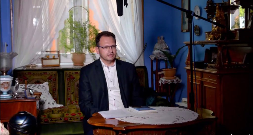 mężczyzna przy stole w pokoju urządzonym w dawnym stylu, u góry widoczny mikrofon ekipy filmowej