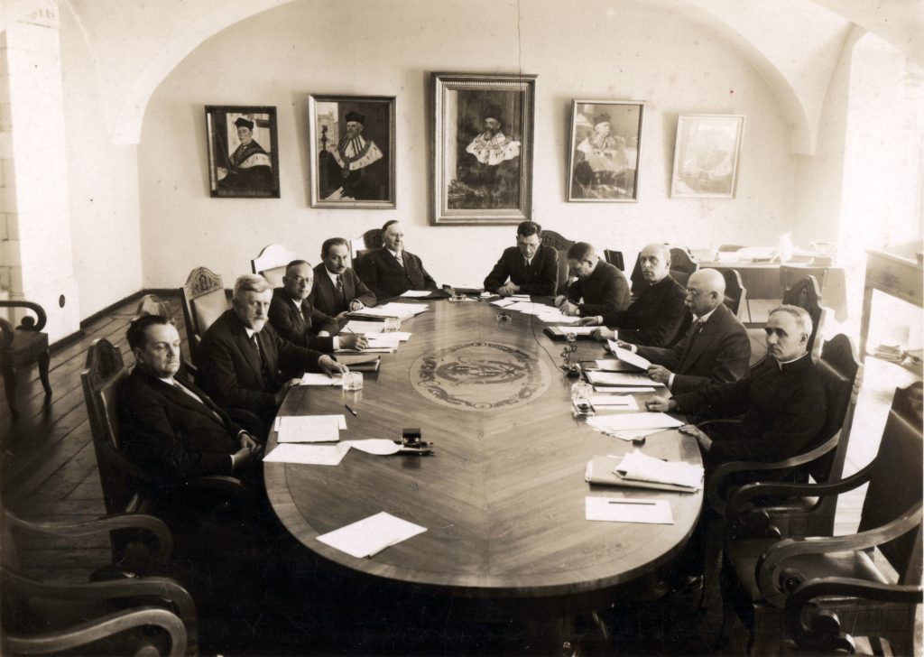 elegancko ubrani mężczyźni siedzący wokół eliptycznego stołu, na ścianie portrety rektorów