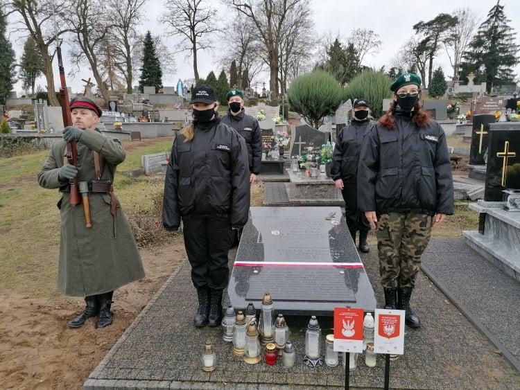grób na cmentarzu. przed nim dwie tabliczki, jedna poświęcona bohaterowi wojny polsko-polszewickiej, druga weteranowi powstania wielkopolskiego. Wokół czwórka młodych ludzi stojących na baczność. Po lewej rekonstruktor salutujący bronią.
