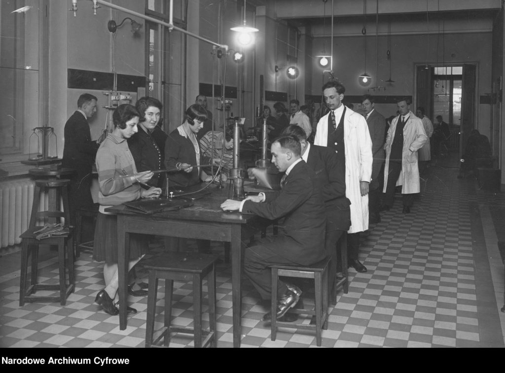 UCZNIOWIE W SALI ZAJĘCIOWEJ, PRZY STOŁACH LABORATORYJNYCH, OBOK INSTRUKTOR W FARTUCHU. zDJĘCIE CZARNO-BIAŁE