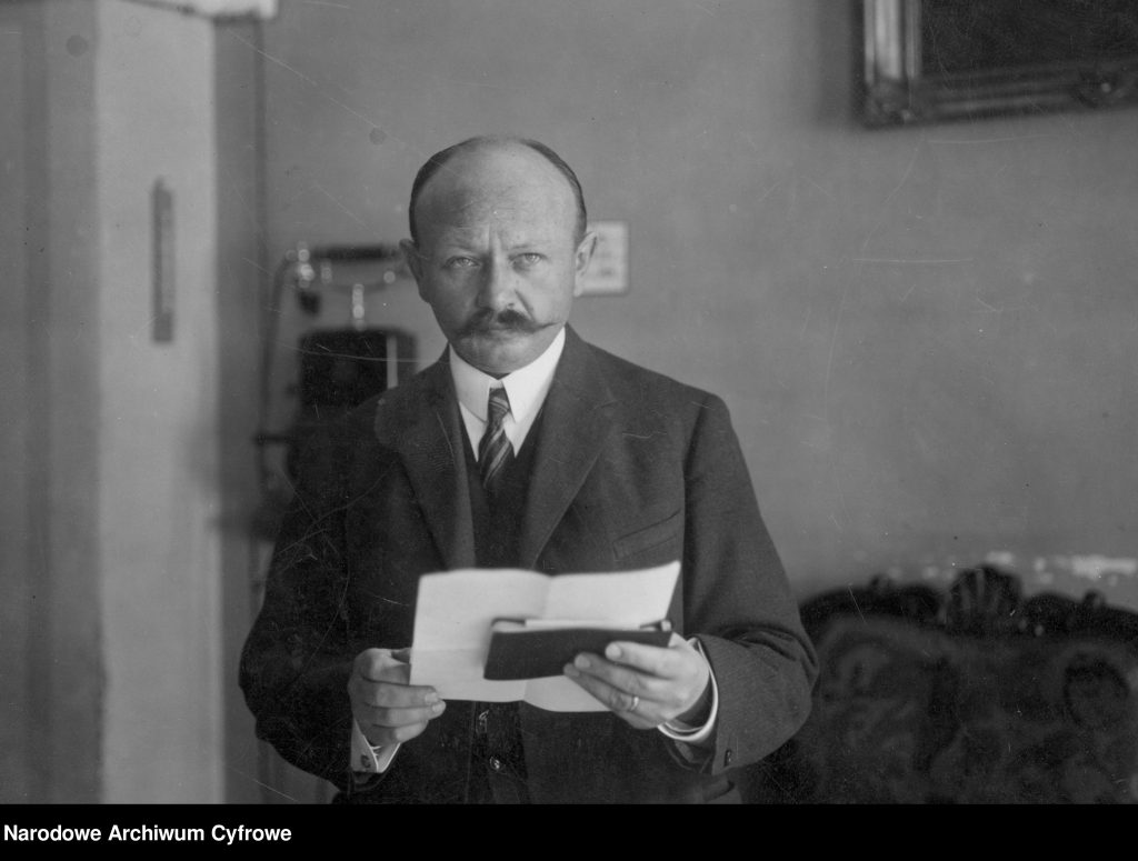 mężczyzna z dokumentami w dłoni w gabinecie naukowym, w tle np. widoczny telefon. Zdjęcie czarno-białe.