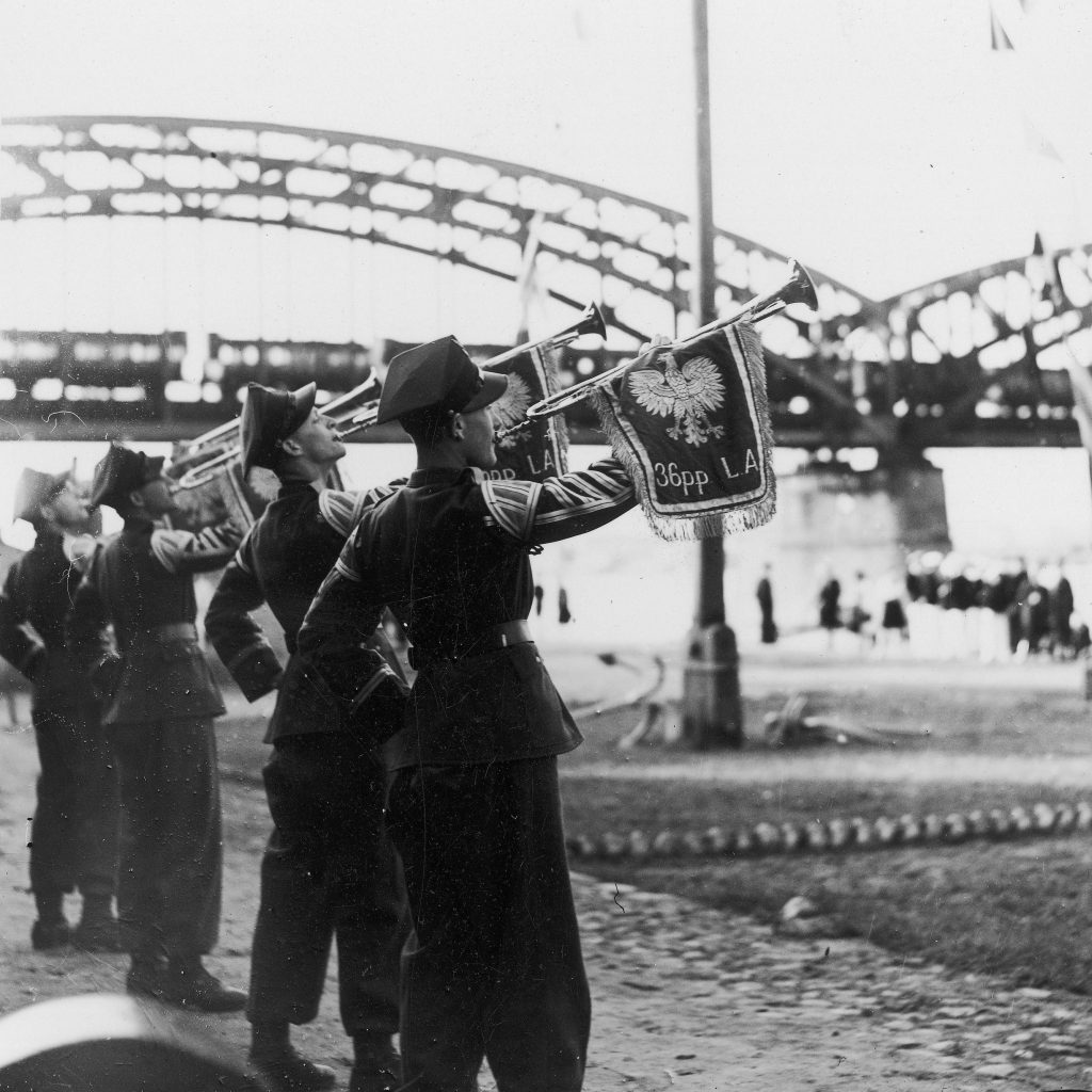 żołnierze w mundurach na brzegu rzeki dmący w trąbki, w tle mosty