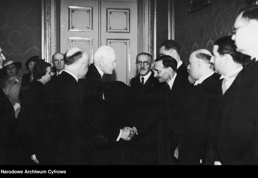 grupa mężczyzny ubranych elegancko wita się ze sobą, w tle drzwi. Zdjęcie czarno-białe.